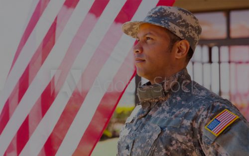 an image of a soldier standing outside on a bright day, looking pensively off to the left with a translucent flag waving behind him